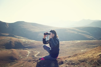 woman wearing black jacket sitting on gray rock and using binoculrs