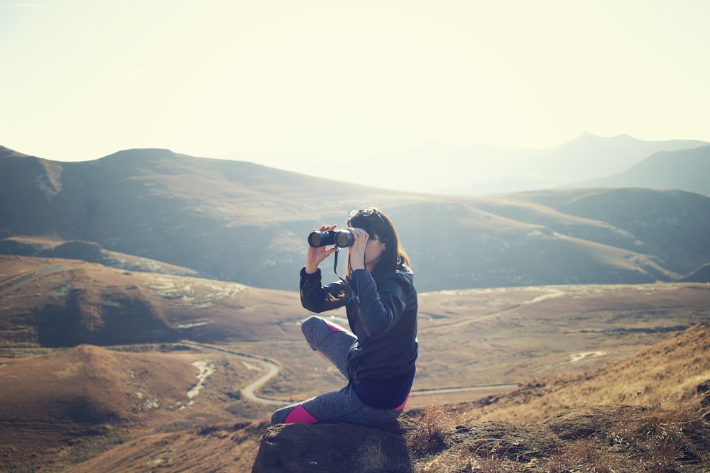 woman wearing black jacket sitting on gray rock and using binoculrs