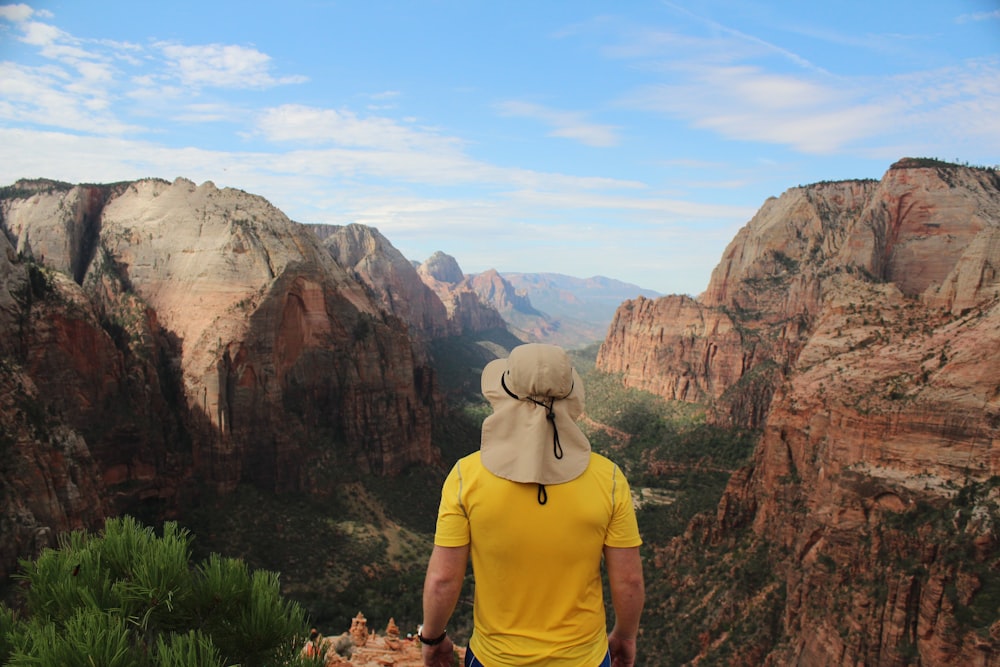 man wearing yellow shirt standing on edge of cliff facing rock formations