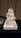 A four-tier white wedding cake decorated with intricate lace-like designs, pink and white roses, and small blue-green flowers. The cake is topped with a figurine of a couple in wedding attire. The tiers are accentuated with rhinestone brooches and pink ribbon bands. It is placed on a decorative white square base on a black tablecloth.