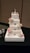 A four-tier white wedding cake decorated with intricate lace-like designs, pink and white roses, and small blue-green flowers. The cake is topped with a figurine of a couple in wedding attire. The tiers are accentuated with rhinestone brooches and pink ribbon bands. It is placed on a decorative white square base on a black tablecloth.
