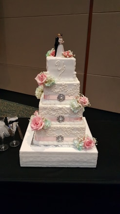 A four-tier white wedding cake decorated with intricate lace-like designs, pink and white roses, and small blue-green flowers. The cake is topped with a figurine of a couple in wedding attire. The tiers are accentuated with rhinestone brooches and pink ribbon bands. It is placed on a decorative white square base on a black tablecloth.