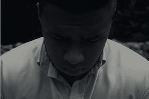 A person wearing a button-up shirt looks downward, casting a shadowed and introspective expression. The scene is captured in a low-light setting, creating a somber and reflective mood.