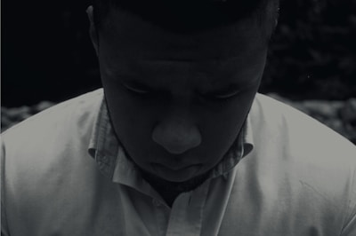 A person wearing a button-up shirt looks downward, casting a shadowed and introspective expression. The scene is captured in a low-light setting, creating a somber and reflective mood.