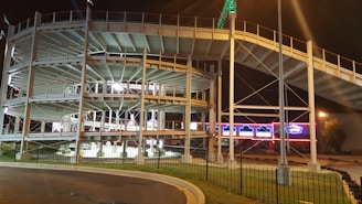 A multi-level go-kart racing track with metal railings, lit by artificial lights at night. Bright signs and lights in the background enhance the vibrant atmosphere.