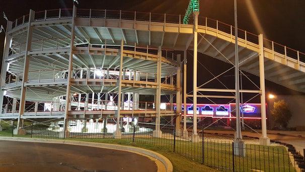 A multi-level go-kart racing track with metal railings, lit by artificial lights at night. Bright signs and lights in the background enhance the vibrant atmosphere.