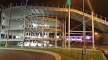A multi-level go-kart racing track with metal railings, lit by artificial lights at night. Bright signs and lights in the background enhance the vibrant atmosphere.