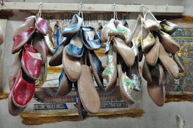 Several pairs of traditional leather shoes hang from hooks against a wall decorated with Egyptian-style art. The shoes feature various colors such as red, black, and metallic designs.