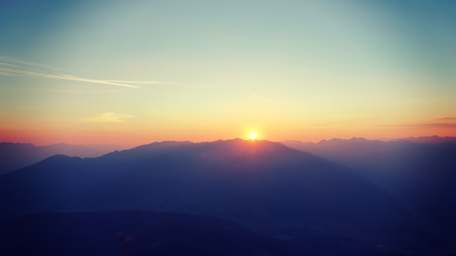 A serene sunset over a mountain range, with the sun setting just above the silhouetted peaks. The sky is a gradient of soft colors from blue to orange, creating a peaceful atmosphere.