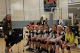 A group of young female volleyball players, all wearing their uniforms, are sitting on a bench, smiling and appearing relaxed. A woman stands to the side, also smiling. The setting is an indoor volleyball court with a net in the background and a scoreboard showing a tied score of 4 to 4.