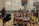 A group of young female volleyball players, all wearing their uniforms, are sitting on a bench, smiling and appearing relaxed. A woman stands to the side, also smiling. The setting is an indoor volleyball court with a net in the background and a scoreboard showing a tied score of 4 to 4.