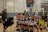A group of young female volleyball players, all wearing their uniforms, are sitting on a bench, smiling and appearing relaxed. A woman stands to the side, also smiling. The setting is an indoor volleyball court with a net in the background and a scoreboard showing a tied score of 4 to 4.