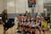 A group of young female volleyball players, all wearing their uniforms, are sitting on a bench, smiling and appearing relaxed. A woman stands to the side, also smiling. The setting is an indoor volleyball court with a net in the background and a scoreboard showing a tied score of 4 to 4.