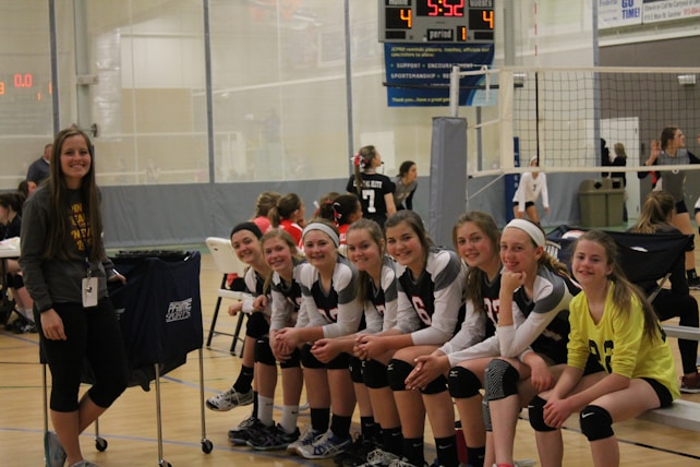 A group of young female volleyball players, all wearing their uniforms, are sitting on a bench, smiling and appearing relaxed. A woman stands to the side, also smiling. The setting is an indoor volleyball court with a net in the background and a scoreboard showing a tied score of 4 to 4.