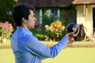 A person wearing a blue shirt is holding a lawn bowl in one hand, seemingly preparing to play a game of lawn bowls. The background features a garden with colorful flowers and a house, giving a pleasant and relaxed outdoor atmosphere.