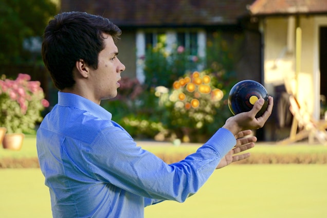 A person wearing a blue shirt is holding a lawn bowl in one hand, seemingly preparing to play a game of lawn bowls. The background features a garden with colorful flowers and a house, giving a pleasant and relaxed outdoor atmosphere.