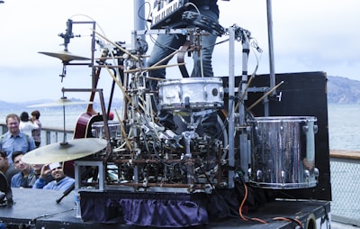 An intricate setup of a street musician's percussion and electronic instrument rig, featuring various drums, cymbals, and wires. The rig appears to be a homemade contraption with a complex arrangement of metal bars and mechanical components. Several onlookers gather around, watching the performance with interest. The backdrop includes a body of water and distant hills under a cloudy sky.