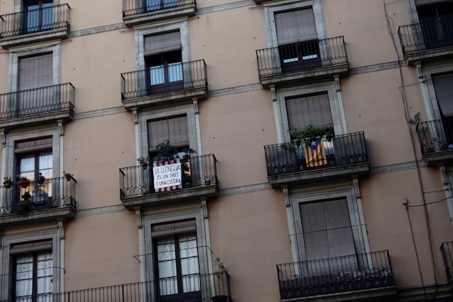 An urban building facade featuring multiple balconies with closed shutters. Two specific balconies stand out: one displaying a sign with text advocating for linguistic rights, and another showing a flag with red and yellow stripes and a blue triangle with a star. Several plants add greenery to the scene.