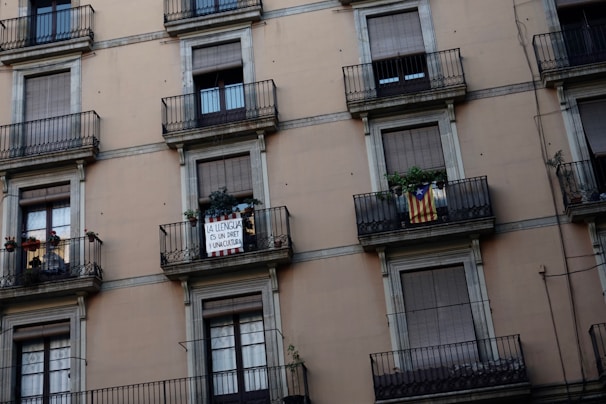 An urban building facade featuring multiple balconies with closed shutters. Two specific balconies stand out: one displaying a sign with text advocating for linguistic rights, and another showing a flag with red and yellow stripes and a blue triangle with a star. Several plants add greenery to the scene.
