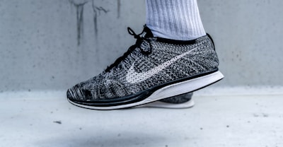 A close-up view of a black and white athletic shoe with a prominent swoosh logo on the side. The shoe is being worn with a white athletic sock, and it appears to be in a mid-air position against a light grey background.