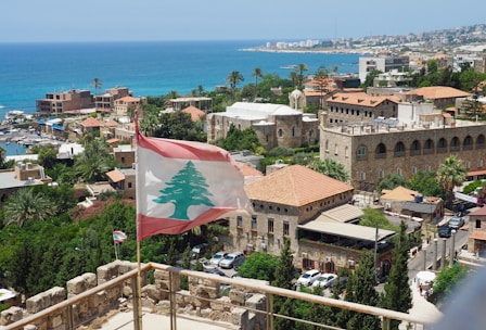 A coastal cityscape featuring a Lebanese flag prominently waving in the foreground. Historical stone buildings with red-tiled roofs are surrounded by lush greenery. The Mediterranean Sea stretches out in the background, accompanied by a clear blue sky.