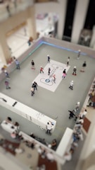 A group of people are roller skating in a designated skating area within a mall. The scene is viewed from above, revealing a rectangular skating rink with a white graphic design in the center. Several skaters of various ages are moving across the grey surface. The surrounding space features spectators and a counter that appears to be offering rental services or assistance.
