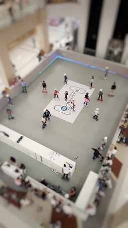 A group of people are roller skating in a designated skating area within a mall. The scene is viewed from above, revealing a rectangular skating rink with a white graphic design in the center. Several skaters of various ages are moving across the grey surface. The surrounding space features spectators and a counter that appears to be offering rental services or assistance.