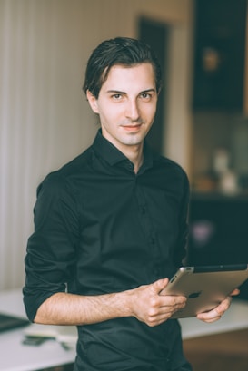 A person wearing a black shirt is holding a tablet in a softly lit indoor setting with a blurred background. The individual has a slight smile and is positioned with one hand holding the tablet as if casually presenting or ready to use it.