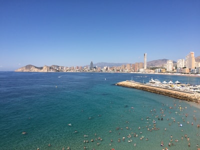 A bustling beach scene with numerous people swimming in clear turquoise waters. The coastline is lined with a cityscape featuring tall modern buildings and mountains in the background. The sky is clear and blue, contributing to the vibrant and lively atmosphere.