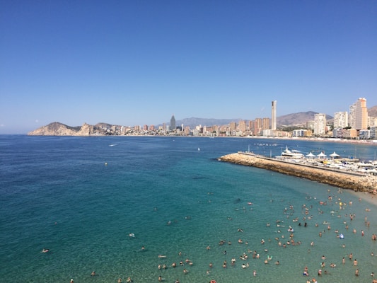 A bustling beach scene with numerous people swimming in clear turquoise waters. The coastline is lined with a cityscape featuring tall modern buildings and mountains in the background. The sky is clear and blue, contributing to the vibrant and lively atmosphere.