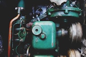A close-up view of a vintage mechanical engine with prominently visible green metal components. The image features various parts including pipes, bolts, and a large cylinder marked 'Oil'. The metal surfaces display signs of rust and wear, highlighting their age and use.