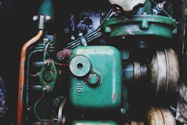 A close-up view of a vintage mechanical engine with prominently visible green metal components. The image features various parts including pipes, bolts, and a large cylinder marked 'Oil'. The metal surfaces display signs of rust and wear, highlighting their age and use.