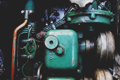 A close-up view of a vintage mechanical engine with prominently visible green metal components. The image features various parts including pipes, bolts, and a large cylinder marked 'Oil'. The metal surfaces display signs of rust and wear, highlighting their age and use.
