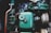 A close-up view of a vintage mechanical engine with prominently visible green metal components. The image features various parts including pipes, bolts, and a large cylinder marked 'Oil'. The metal surfaces display signs of rust and wear, highlighting their age and use.