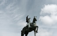 A statue of a warrior on horseback is prominently displayed against a partly cloudy sky. The statue depicts a historical figure with intricate armor, raising one arm commandingly while sitting confidently on a rearing horse.