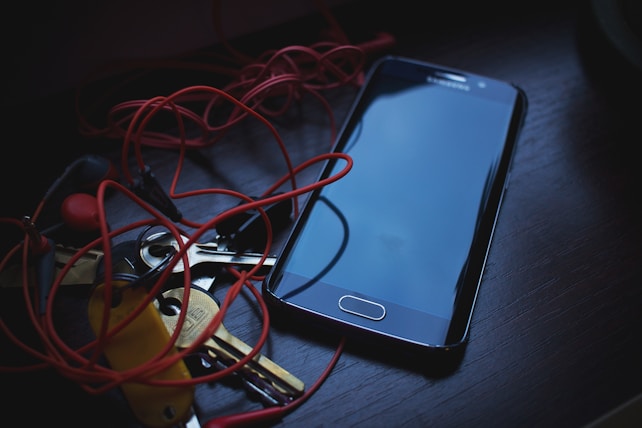 A smartphone is placed on a dark wooden surface surrounded by tangled red earphones and several metallic keys. The phone screen is off and reflective. The scene is dimly lit, casting soft shadows and giving it a moody atmosphere.