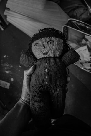 A hand holding a knitted doll with button-like eyes and a simple stitched face. The doll has textured clothing and yarn hair, with a worn appearance. The background includes blurry, indistinct items, possibly boxes or papers, creating a cluttered and nostalgic atmosphere.