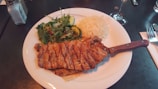 A plate containing grilled steak with grill marks, accompanied by a fresh salad with cucumbers and greens, and a serving of white rice. Silverware and a napkin are placed nearby, with a glass of water or wine partially visible.