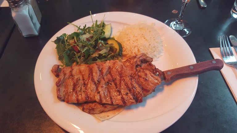 A plate containing grilled steak with grill marks, accompanied by a fresh salad with cucumbers and greens, and a serving of white rice. Silverware and a napkin are placed nearby, with a glass of water or wine partially visible.