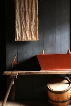 A rustic setting with a wooden bench holding a red, angled trough. Behind it is a dark wooden panel wall, from which a beige cloth hangs vertically. Below, a round wooden barrel is partially visible beneath the bench.