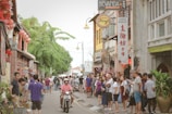 A bustling street scene in an urban area with people walking and standing along the sidewalk. A man rides a motorbike down the street, and there are various shops with signs, including one for 'Kedai Biskut'. The street is lively, with vibrant plants and traditional decorations, including lanterns.