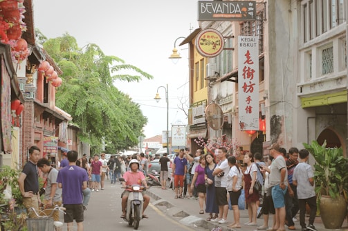A bustling street scene in an urban area with people walking and standing along the sidewalk. A man rides a motorbike down the street, and there are various shops with signs, including one for 'Kedai Biskut'. The street is lively, with vibrant plants and traditional decorations, including lanterns.