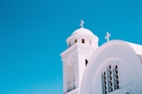 A white church with arched windows and two prominent crosses, set against a bright blue sky. The structure features a dome and architectural elements typical of Mediterranean-style churches.