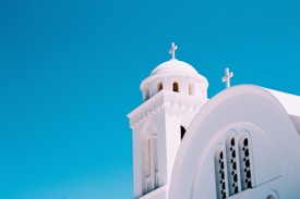 A white church with arched windows and two prominent crosses, set against a bright blue sky. The structure features a dome and architectural elements typical of Mediterranean-style churches.