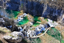 A picturesque landscape features cascading waterfalls flowing into vivid turquoise pools, surrounded by rocky cliffs and sparse vegetation. A wooden pathway meanders through the water, offering a scenic route across the scene.