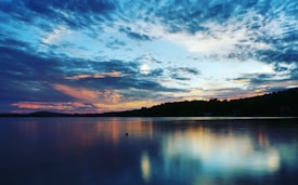 The image features a serene lake during sunset, where the sky is filled with dramatic clouds in shades of blue, orange, and pink. The reflection of the sky creates a mirror-like effect on the still water, enhancing the natural beauty.