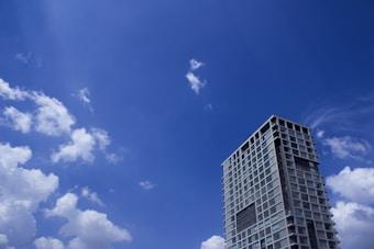 A modern high-rise building with numerous windows stands against a vibrant blue sky dotted with scattered white clouds.