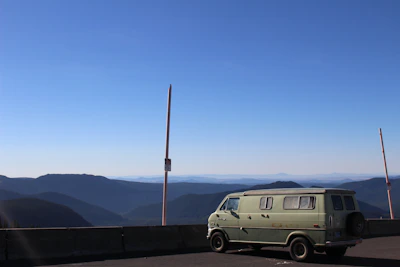 A panoramic shot of the van on a mountain road, clouds rolling beneath the peaks.