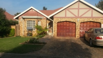 A brick house with a gable roof and two wooden garage doors. The house is surrounded by a well-maintained yard with green grass and some shrubs. A silver car is parked in the driveway.