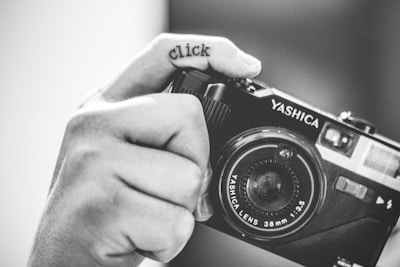 A black and white image featuring a close-up of a hand holding a vintage Yashica camera. The word 'click' is tattooed across the finger, adding an artistic touch to the composition.